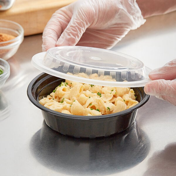 A gloved hand places a clear lid on a round black plastic container filled with pasta.