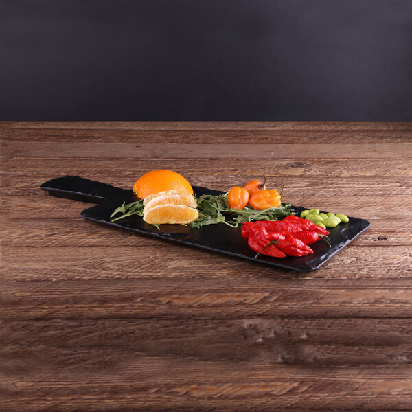 An Elite Global Solutions faux slate serving board with a plate of oranges, peppers, and other vegetables on a wood table.