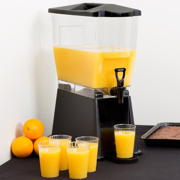 A Carlisle black premium beverage dispenser with a glass of orange juice on a table with four empty glasses.