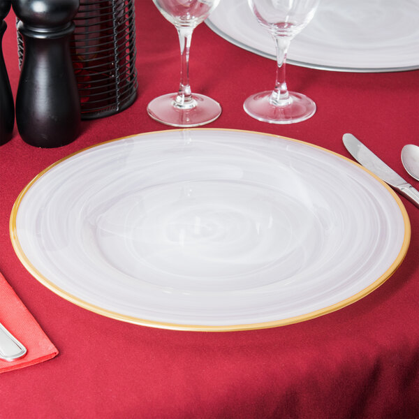 A white Charge It by Jay alabaster glass charger plate with a gold rim on a table with glasses and a knife on a red tablecloth.