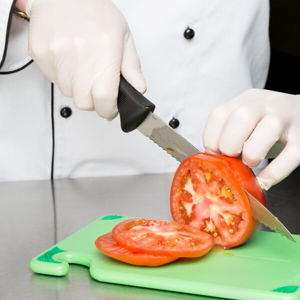 A person in white gloves uses a Mercer Culinary Millennia 8" Serrated Edge Utility Knife to cut a tomato on a cutting board.