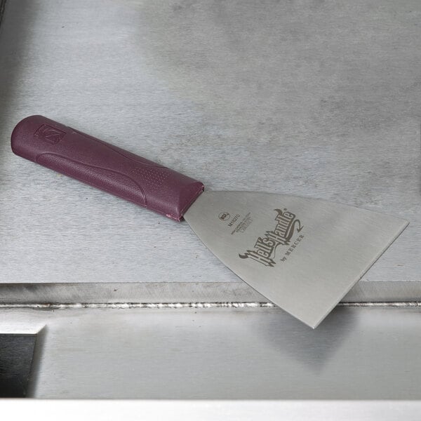 A Mercer Culinary grill scraper with a burgundy handle on a metal surface.