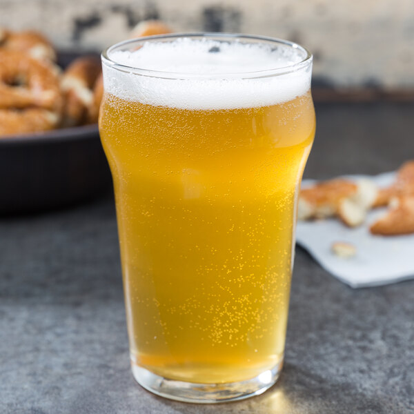 A Libbey English Pub nonic glass of beer with foam next to a bowl of pretzels on a table.