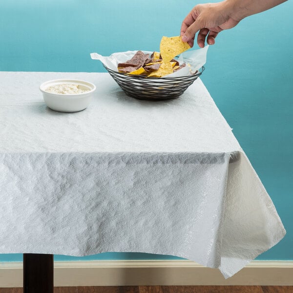 A hand dipping an Atlantis Plastics white pebble embossed plastic table cover into a bowl of chips on a table.