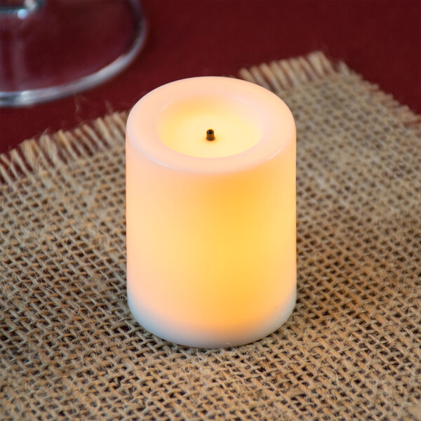 A Sterno white flameless real wax mini votive candle on a burlap surface next to a glass.