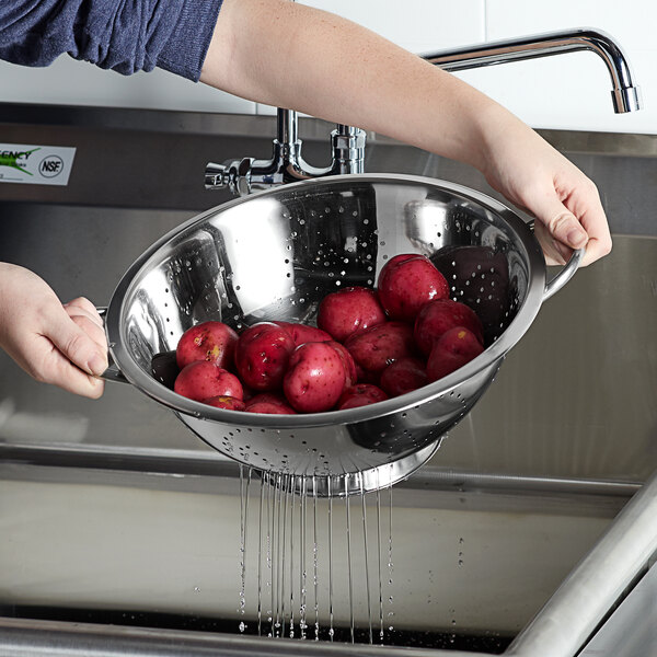 A person washing red apples in a Vollrath stainless steel colander with base and handles.