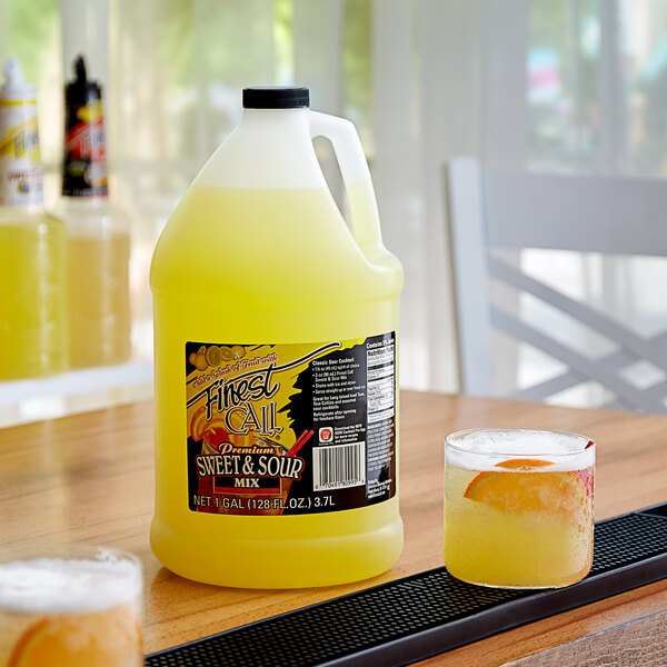 A jug of Finest Call sweet and sour mix next to a glass of yellow liquid.