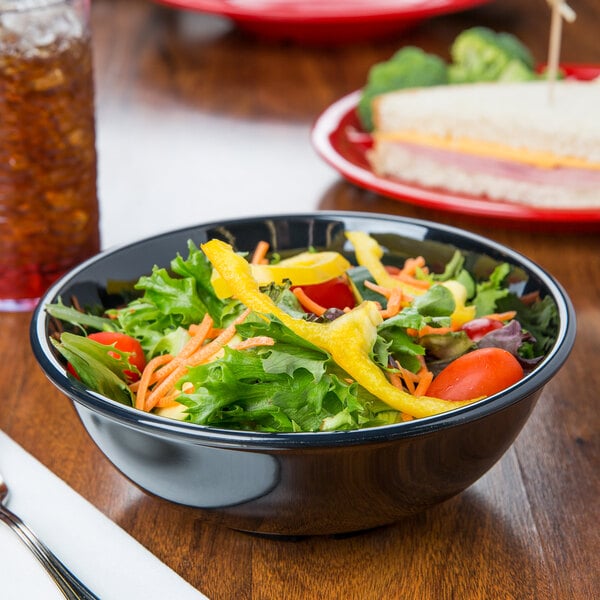A black melamine bowl filled with salad on a table with a sandwich and a drink.