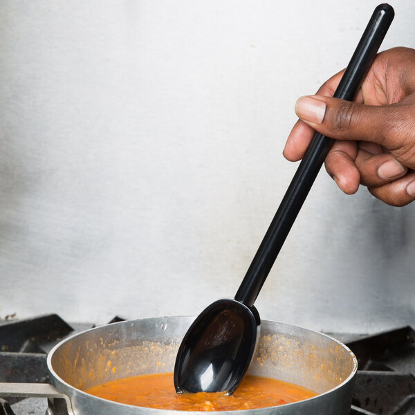 A person holding a Mercer Culinary black high temperature mixing spoon over a pot of soup.