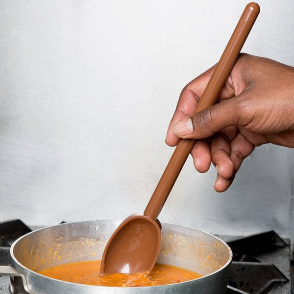 A person stirring soup in a pot with a brown Mercer Culinary Hell's Tools high temperature mixing spoon.