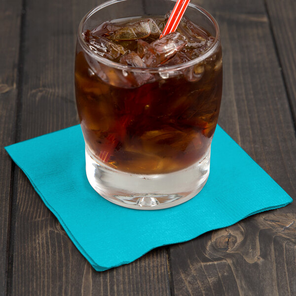 A glass of iced coffee with a Bermuda Blue Creative Converting beverage napkin and a straw on a table.