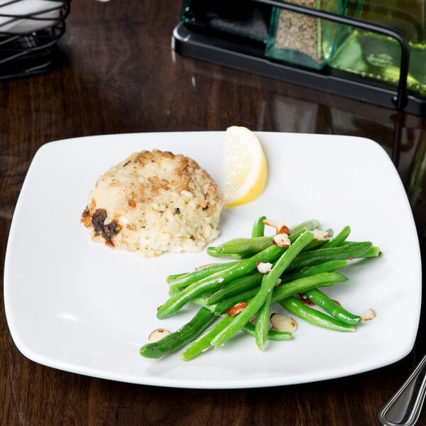 A Libbey square porcelain plate with food, green beans, and a lemon wedge on a table.