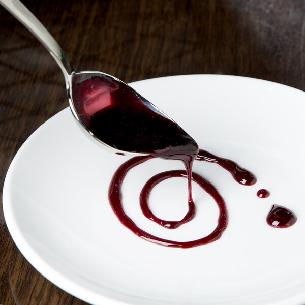 A Mercer Culinary petite saucier spoon with a spoonful of red liquid on a white plate.