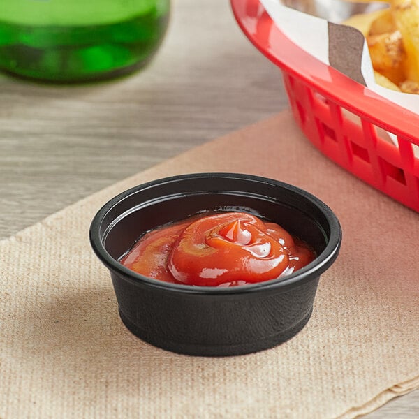 A black plastic souffle cup filled with ketchup next to a bowl of fries.