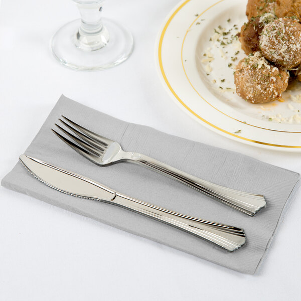 A fork and knife on a Shimmering Silver paper dinner napkin next to a plate of food.
