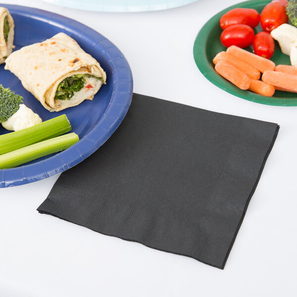 A pack of black Creative Converting luncheon napkins on a table with a plate of vegetables and wraps.