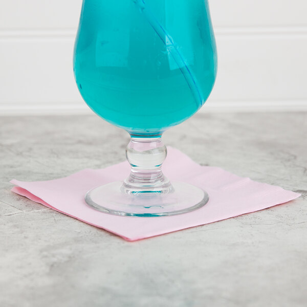 A blue drink in a glass with a pink straw on a table with a Classic Pink beverage napkin.