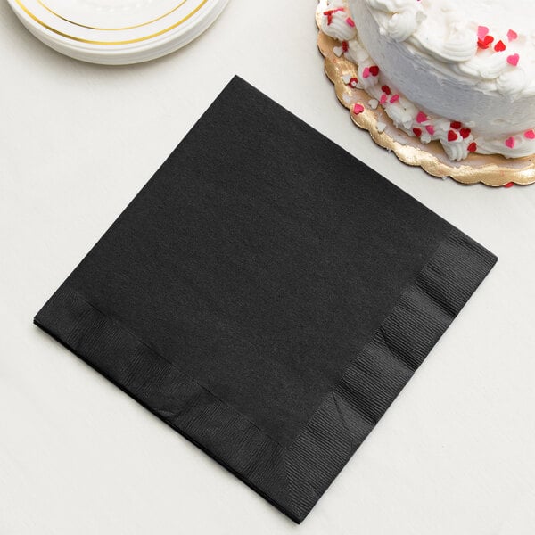 A black Creative Converting paper dinner napkin on a stack of plates with a gold line next to a white cake with red and pink sprinkles.