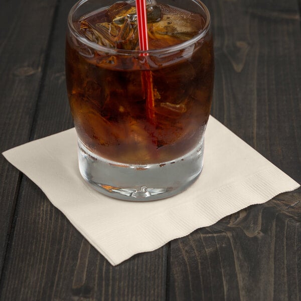 A glass of brown liquid with ice and a Creative Converting ivory beverage napkin with a straw.