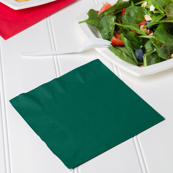 A pack of Hunter Green 1/4 Fold Luncheon Napkins on a table with a plate of salad and a fork.