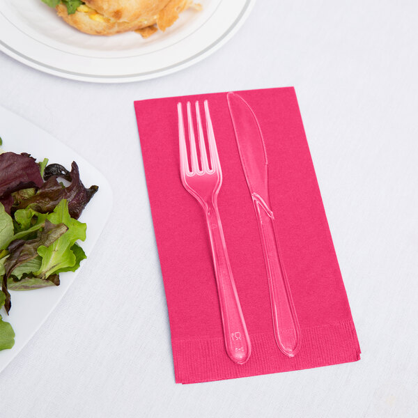 A fork and knife on a hot magenta pink Creative Converting guest towel next to a plate of salad.
