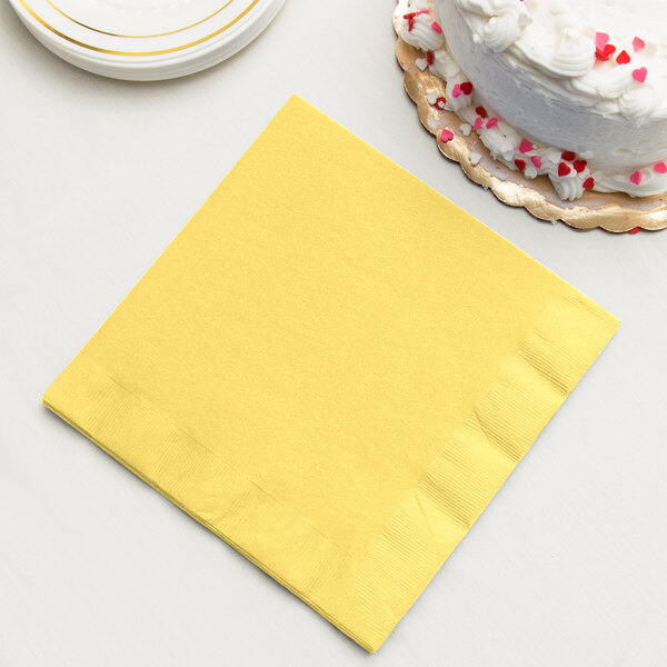 A stack of yellow Creative Converting paper dinner napkins with a slice of white cake on a plate.