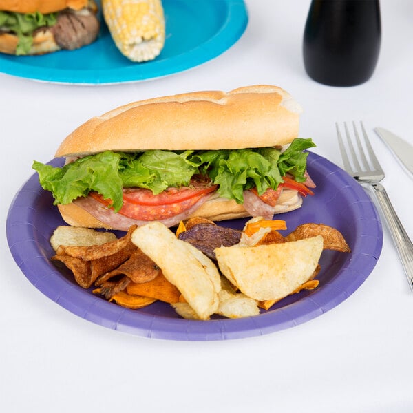 A Creative Converting purple paper plate with a sandwich, chips, and a fork on a table.