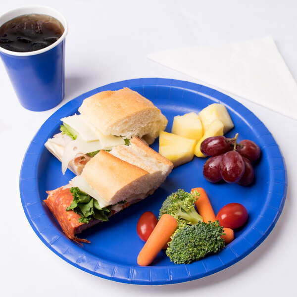 A Creative Converting cobalt blue paper plate with a sandwich, fruit, and a drink on it.