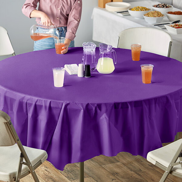 A purple table with a Creative Converting Amethyst Purple OctyRound plastic table cover and a glass of orange juice.