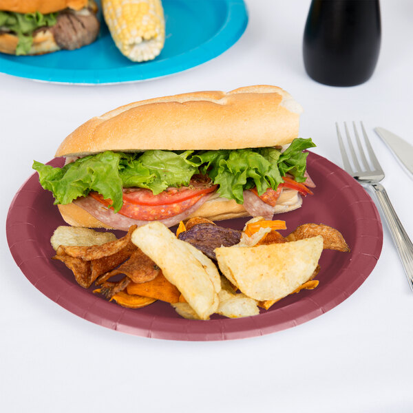 A sandwich with lettuce and tomato on a Creative Converting burgundy paper plate with a fork and knife.