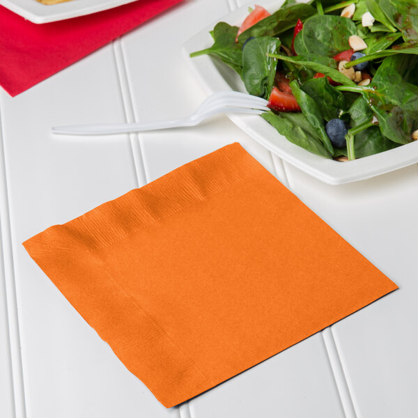 A white plate of salad with orange napkins on the table.