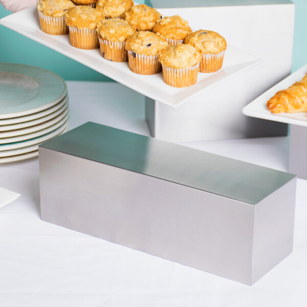 A rectangular stainless steel riser with a tray of muffins on a table.