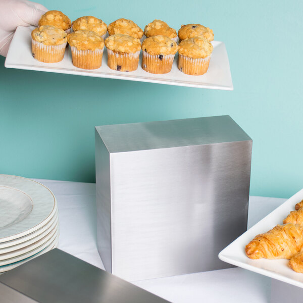 A person holding a plate of muffins on a stainless steel rectangular riser.