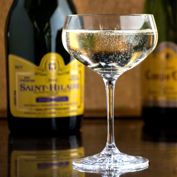 A Spiegelau Perfect Serve coupe glass filled with a bubbly liquid on a table with wine bottles.