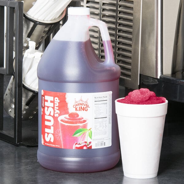 A jug of Carnival King Cherry Slushy Concentrate on a counter next to a white cup of slushy with a red lid.