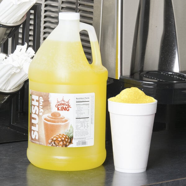 A jug of yellow liquid with a Carnival King label next to a white cup of yellow slushy with a pineapple slice on the rim.