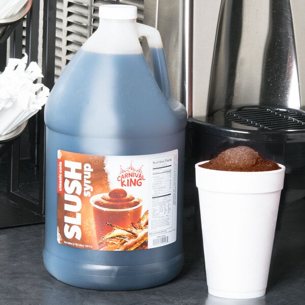 A jug of Carnival King Cola Slushy Concentrate next to a cup of slushy ice.