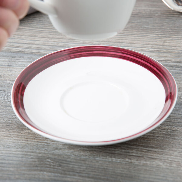 A hand holding a 10 Strawberry Street white porcelain cup over a raspberry rim saucer.