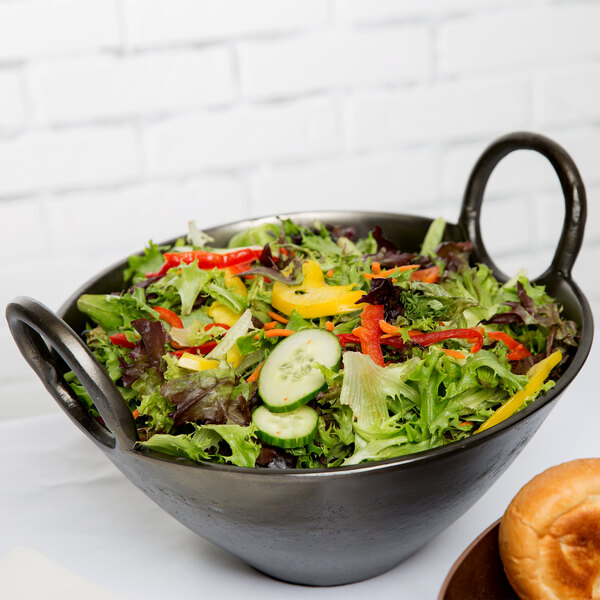 A bowl of salad with a bagel and a plate of bread in a black metal bowl with raw black metal handles.