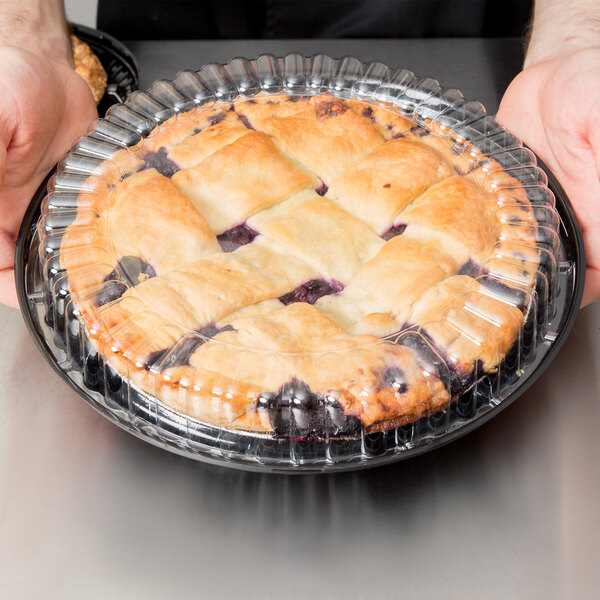 A person holding a pie in a D&W Fine Pack black plastic container with clear low dome lid.