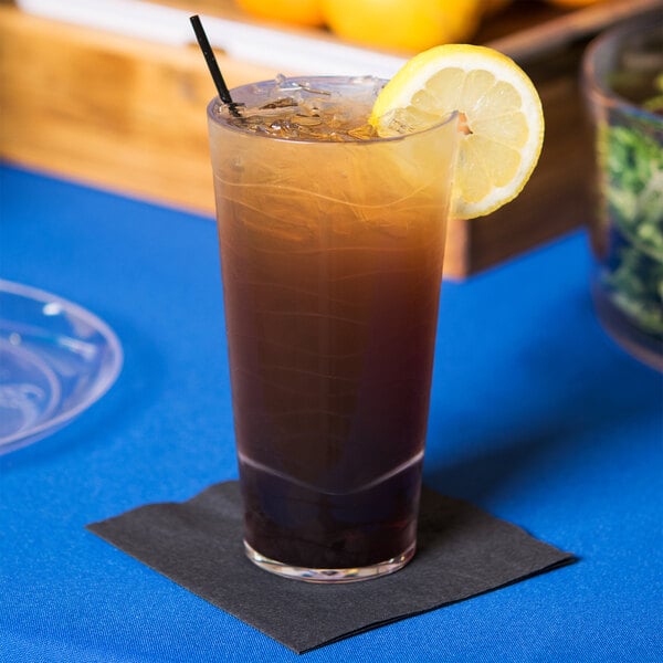 A clear plastic tumbler with a straw and a lemon wedge in a brown drink on a table.