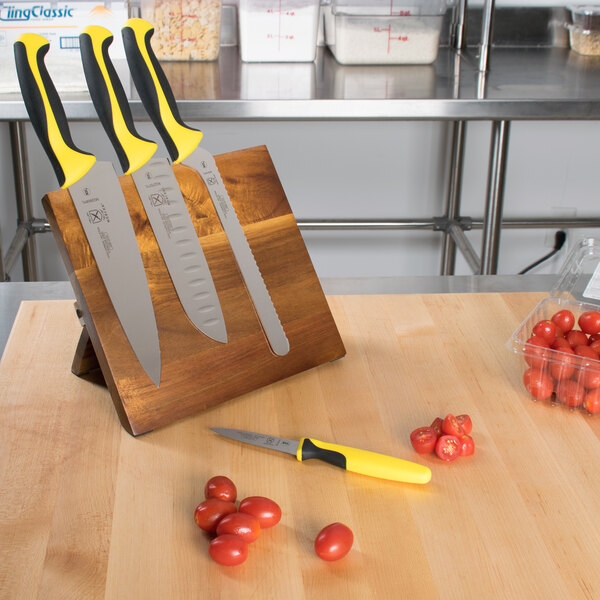 A Mercer Culinary Millennia Colors knife set on an acacia cutting board with yellow and black handles, next to tomatoes.