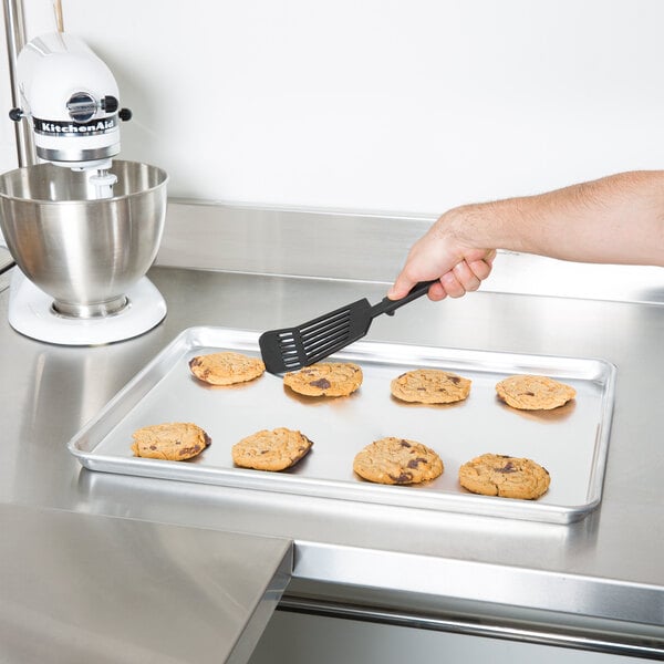 A hand holding a spatula over Vollrath Wear-Ever bun pan of cookies.