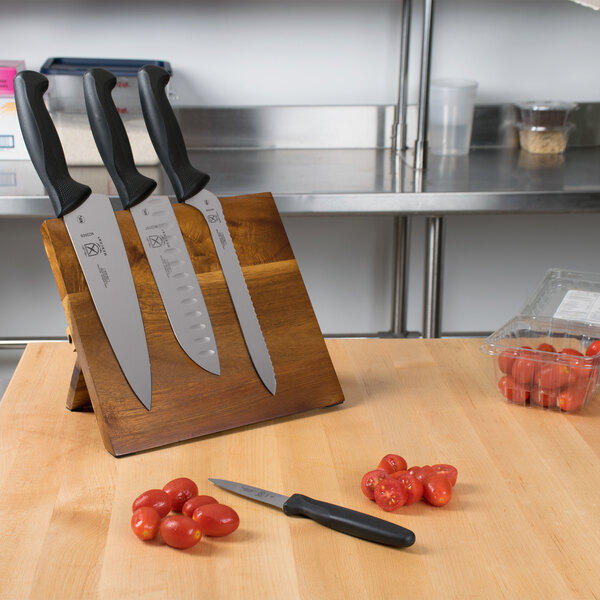 A Mercer Culinary Millennia knife set on an acacia magnetic board with black handles next to tomatoes.