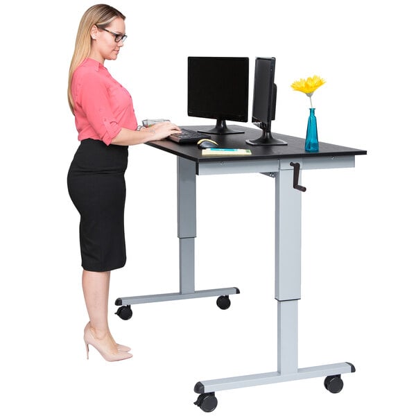A woman standing at a black and gray Luxor stand up desk with a computer.