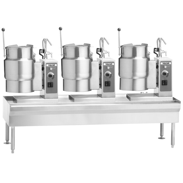 A white rectangular Vulcan table with three electric tilting kettles on it.
