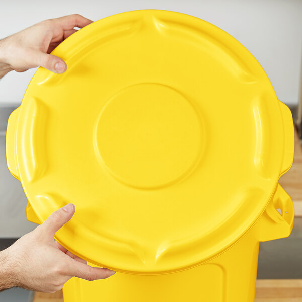 A hand holding a yellow Rubbermaid BRUTE trash can lid.