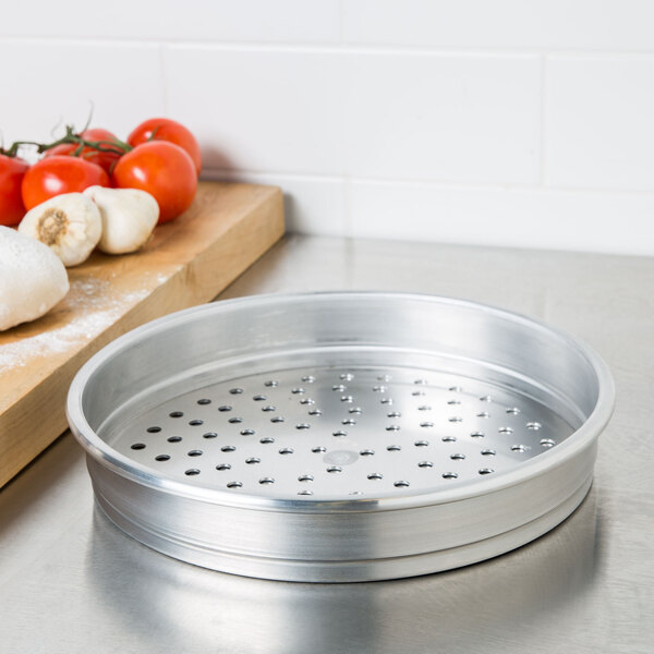 An American Metalcraft heavy weight aluminum pizza pan with holes in it sitting on a counter next to tomatoes and garlic.