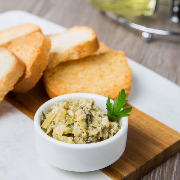 A white porcelain ramekin filled with green pesto on a table with bread and crackers.