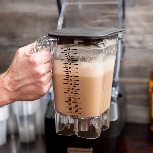 A hand using a Hamilton Beach blender with a brown liquid in a clear container with a lid and blade.